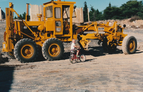 A young boy on a red bicycle posing in front of construction machinery. 
