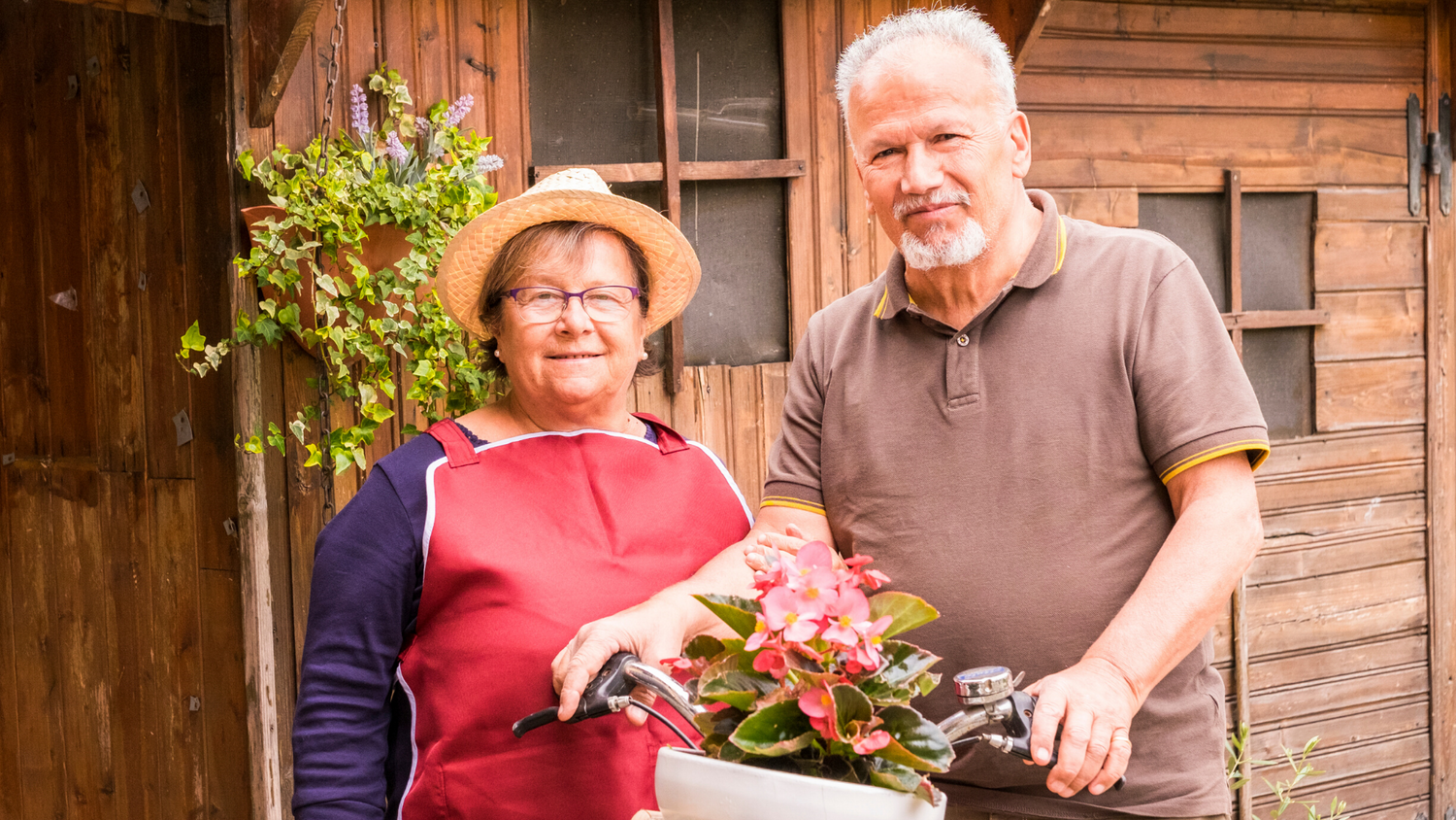 An elderly couple standing in front of their home. The man is holding a potted plant.