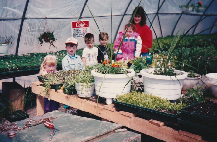 A woman and three young children standing inside a greenhouse, surrounded by plants.